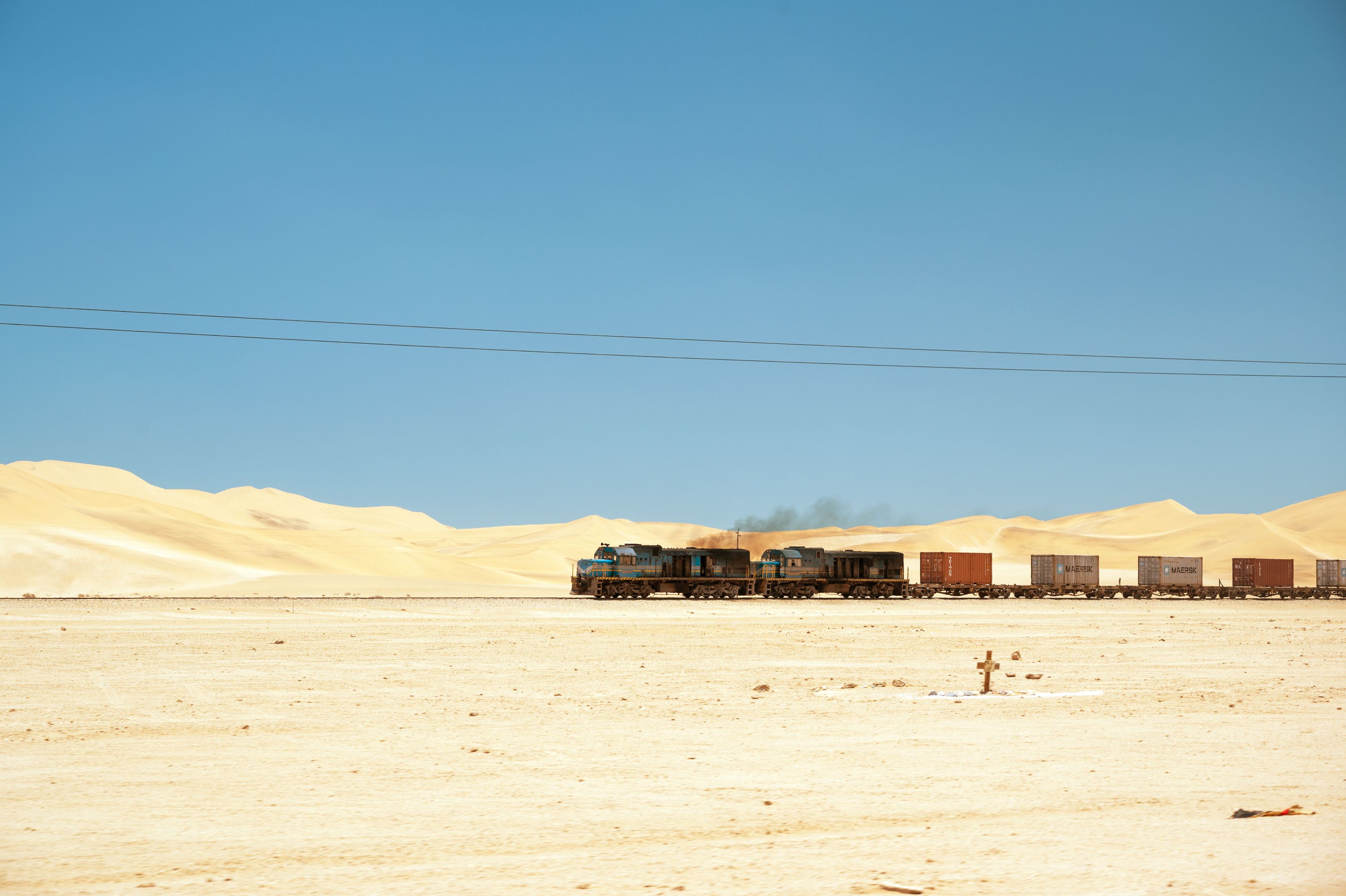 Eine idyllische Wüstenlandschaft mit Dünen wird von Gleisen und Stromleitungen durchbrochen. Man sieht Die Lokomotive und ein paar Wagen eines schweren Güterzugs auf den Gleisen fahren. Aus dem Auspuff der Lokomotive kommt eine schwarze Rauchwolke. Im Vorderung liegt ein einsames Grab mit Kreuz.