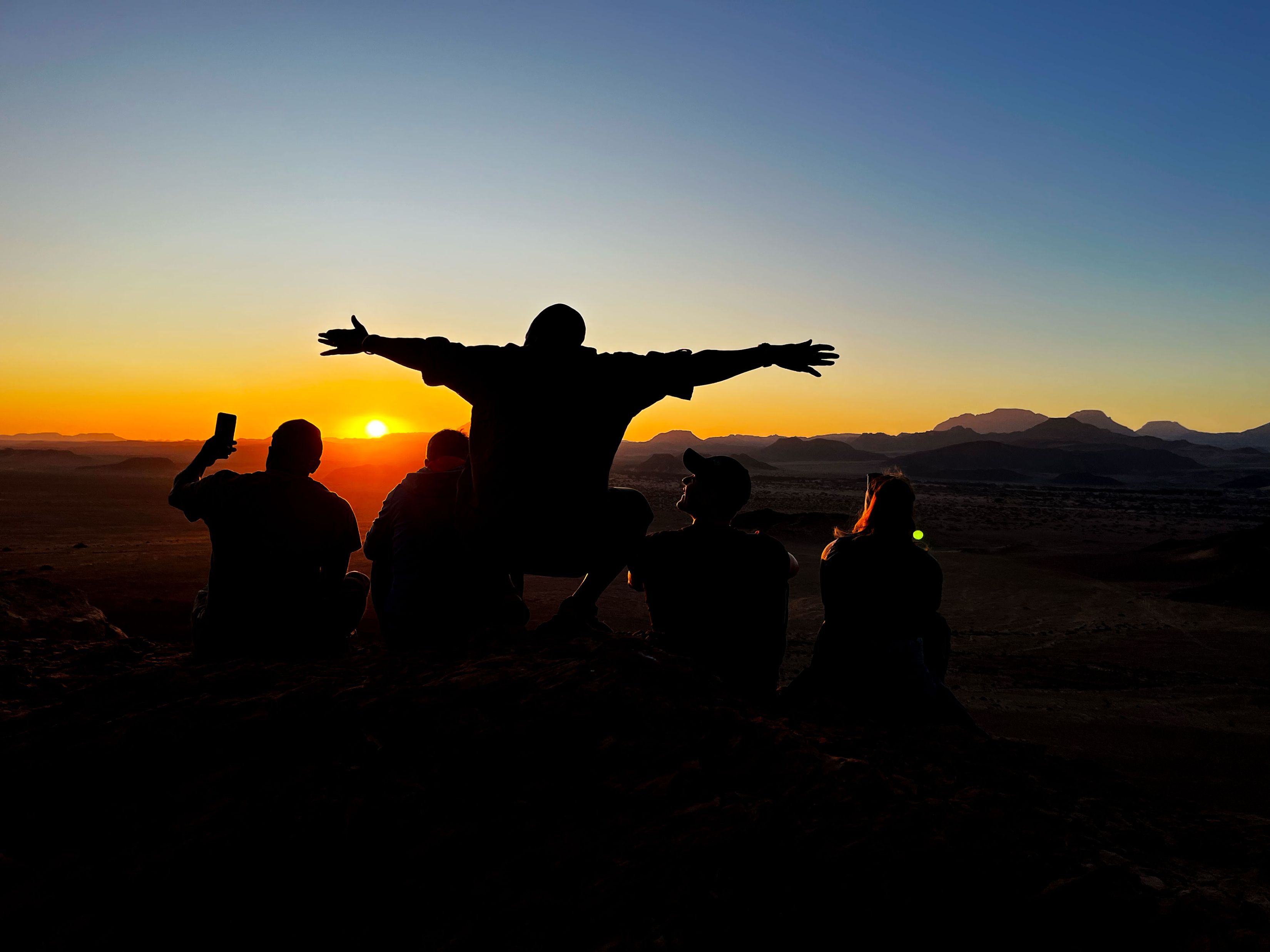 Die Aufnahme ist auf einer Bergkuppe während einem farbenfrohen Sonnenaufgang im Damaraland gemacht worden. Auf der Koppe sieht man vier Menschen von hinten im Gegenlicht: Ein Mann steht und streckt die Arme seitlich aus, ein Mann und eine Frau sitzen rechts von ihm und ein junger Mann, der links sitzt, hält ein Handy in die Höhe. Die Sonne ist knapp über dem Horizont aufgetaucht und taucht den Himmel in gold, orange, gelb und blau.
